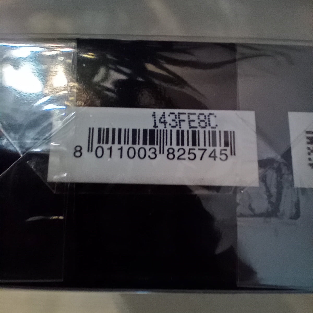 A close-up of a packaged black item inside a clear plastic sleeve, with a white barcode label showing the number 143FE8C and a barcode underneath.
