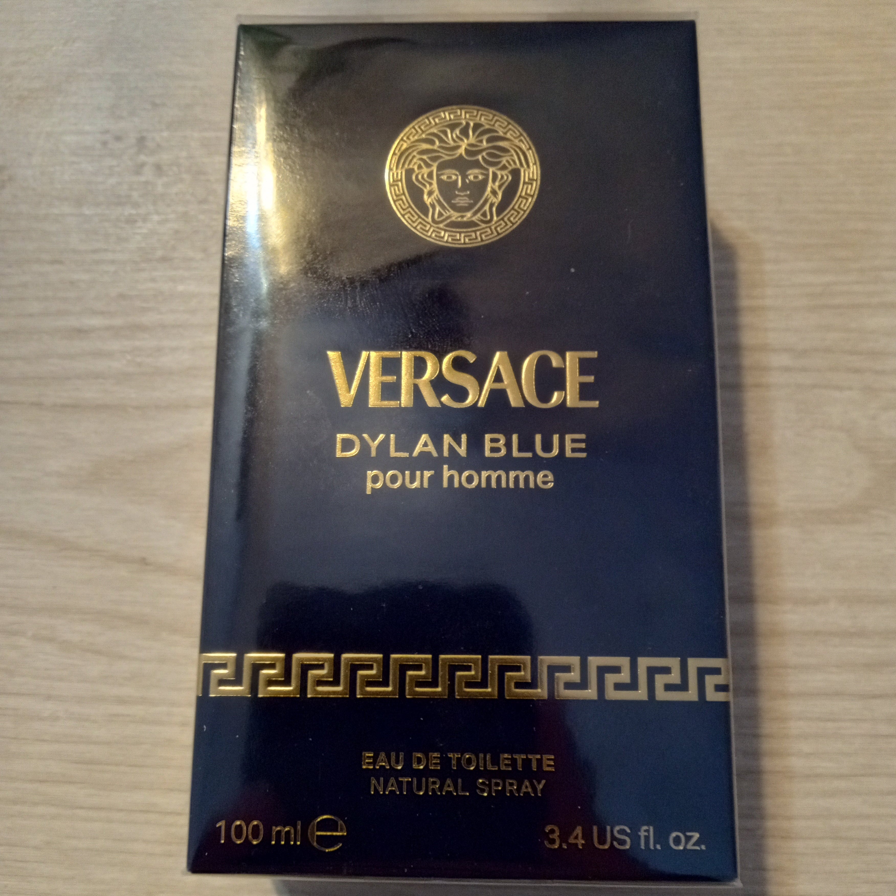 A 100ml box of Versace Dylan Blue pour homme Eau de Toilette, featuring a dark navy blue box with gold lettering and the iconic Medusa logo, placed on a light wood surface.