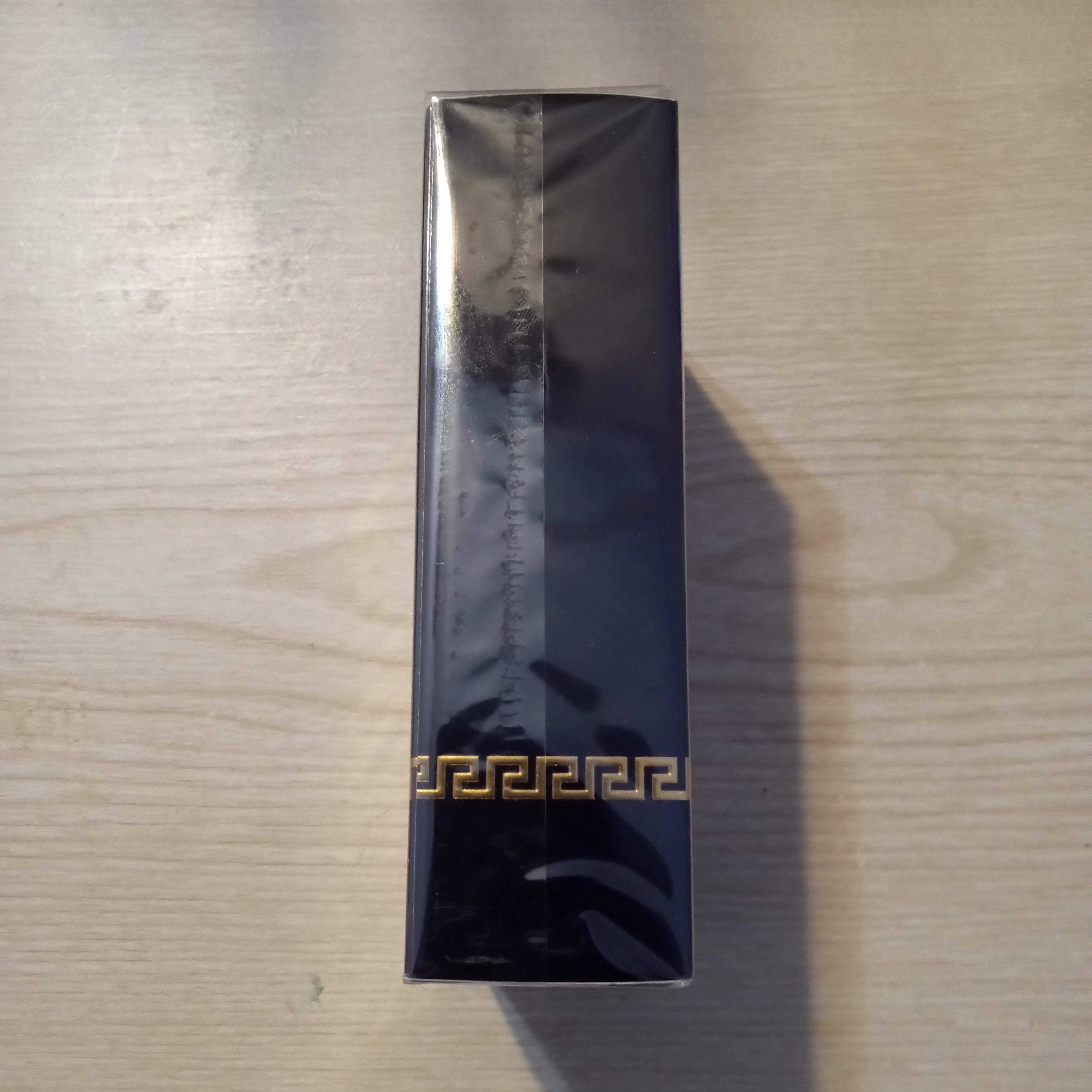 A tall, narrow black rectangular box wrapped in protective plastic, featuring a gold Greek key pattern near the bottom, placed on a light wood surface.