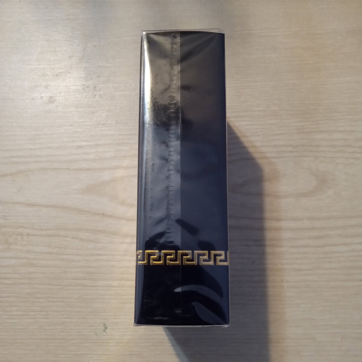 A tall, narrow black rectangular box wrapped in protective plastic, featuring a gold Greek key pattern near the bottom, placed on a light wood surface.