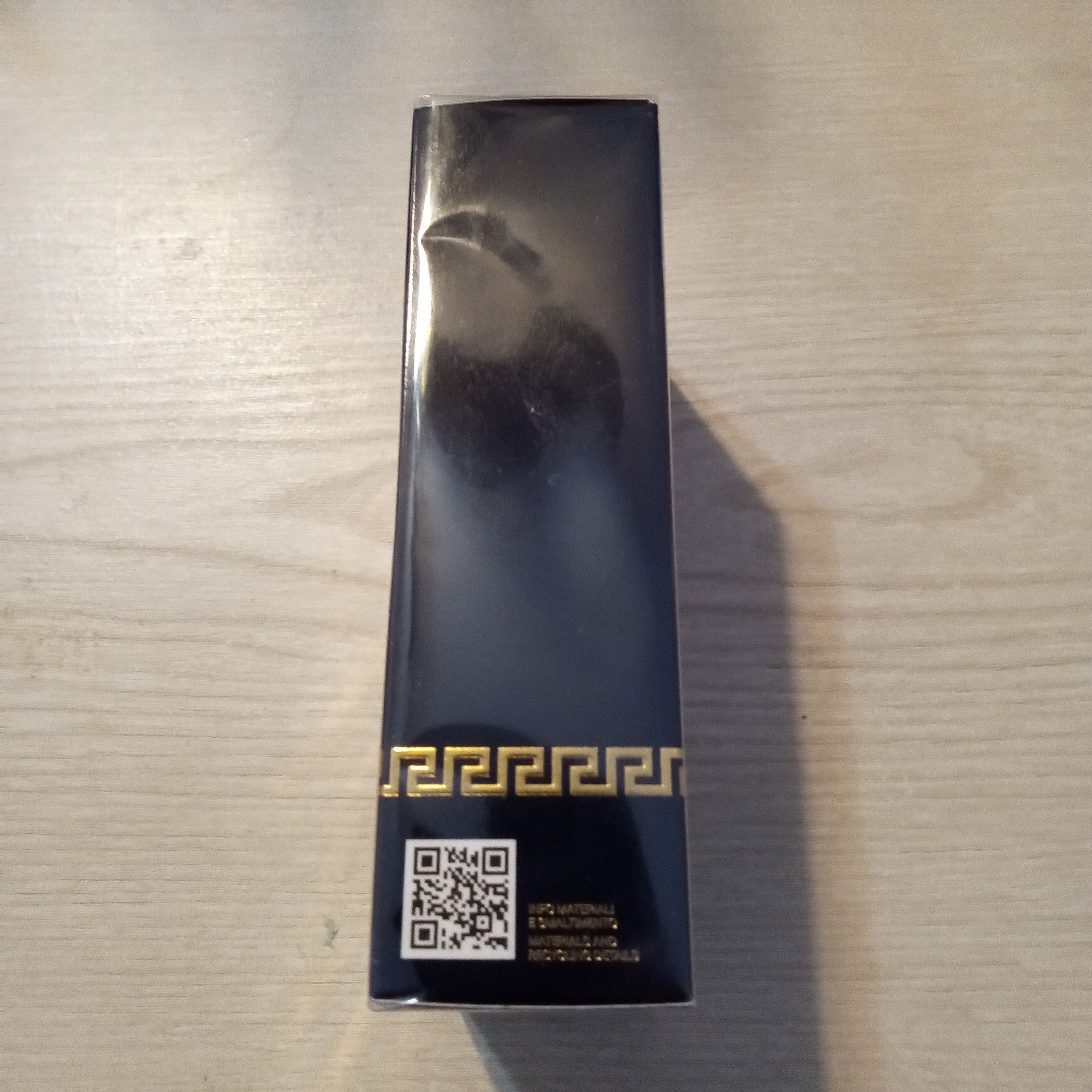 A tall, narrow black product box with a glossy finish, featuring a gold Greek key pattern near the bottom and a QR code, placed on a light wood tabletop.