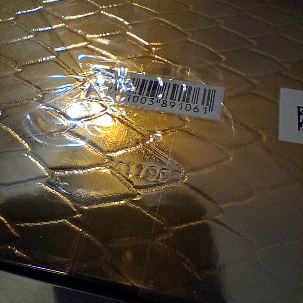 A close-up of a gold textured box with a crocodile or alligator pattern, sealed in clear plastic with a barcode sticker on top.