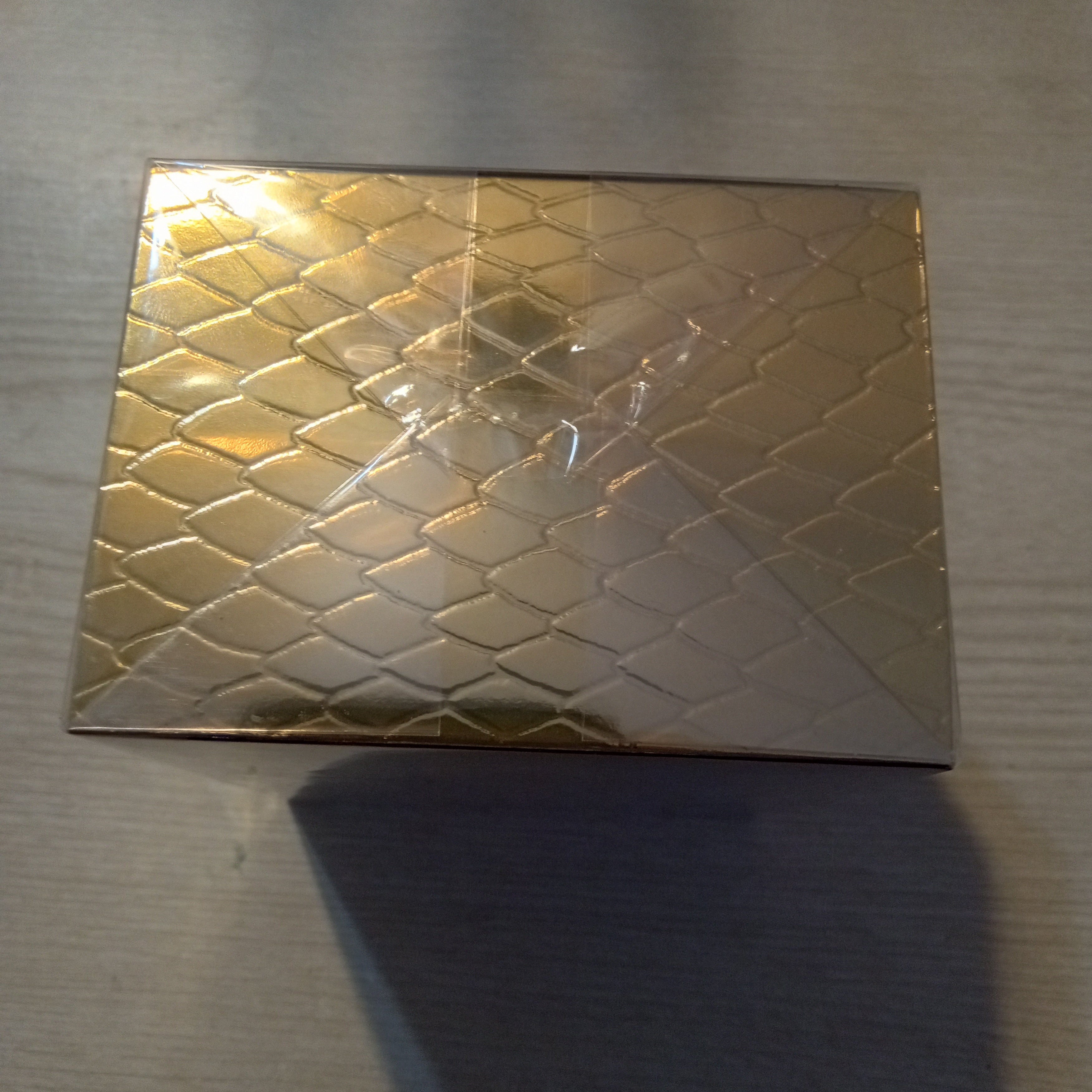 A rectangular gold-toned box with a raised diamond pattern, photographed on a light gray tabletop with a strong shadow underneath.