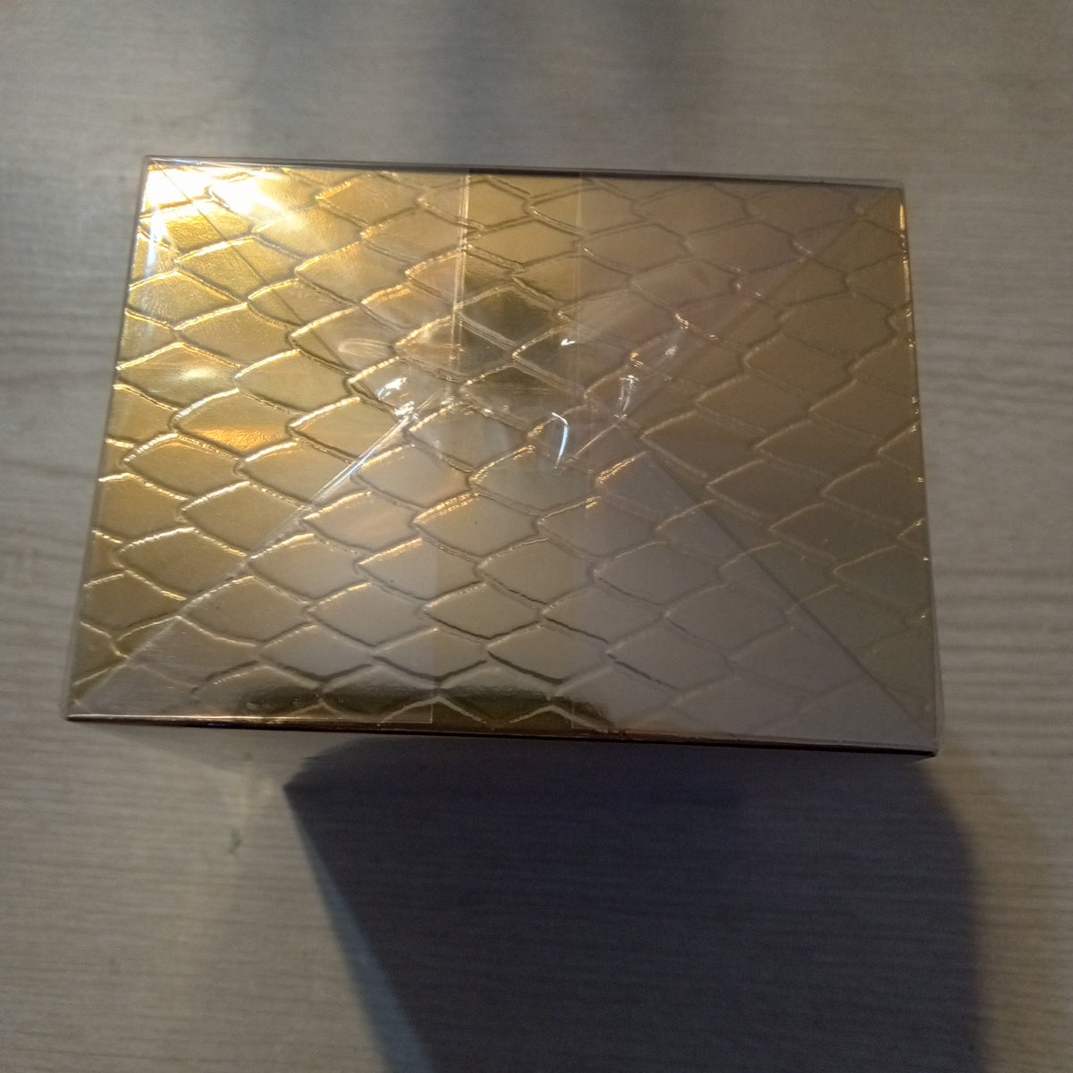 A rectangular gold-toned box with a raised diamond pattern, photographed on a light gray tabletop with a strong shadow underneath.