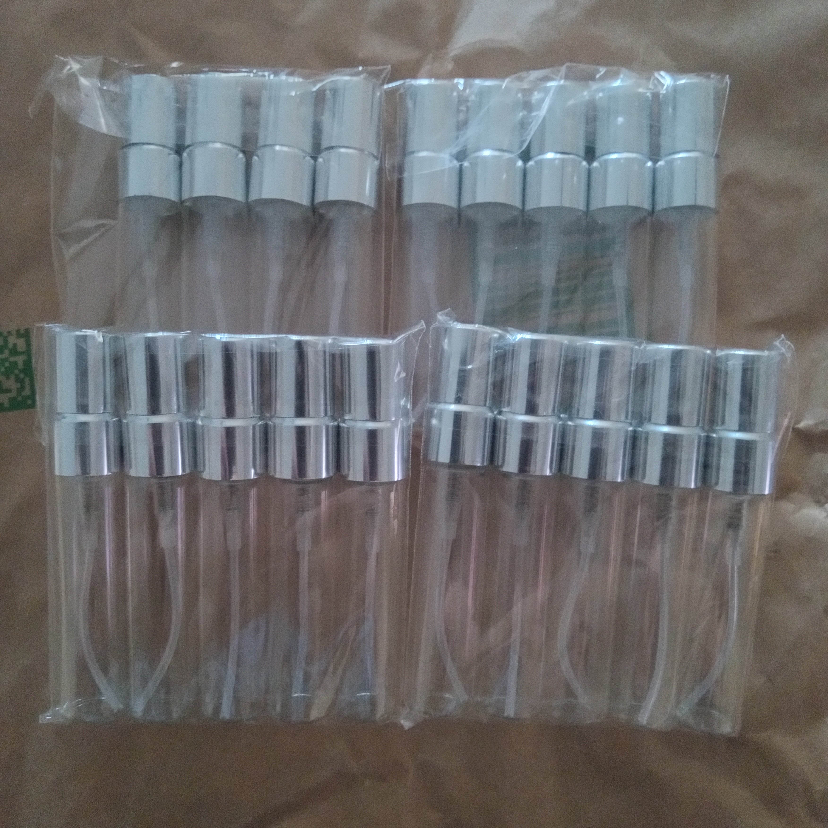 Six clear plastic spray atomizer bottles with silver metal collars, individually wrapped in clear plastic and arranged on brown paper.