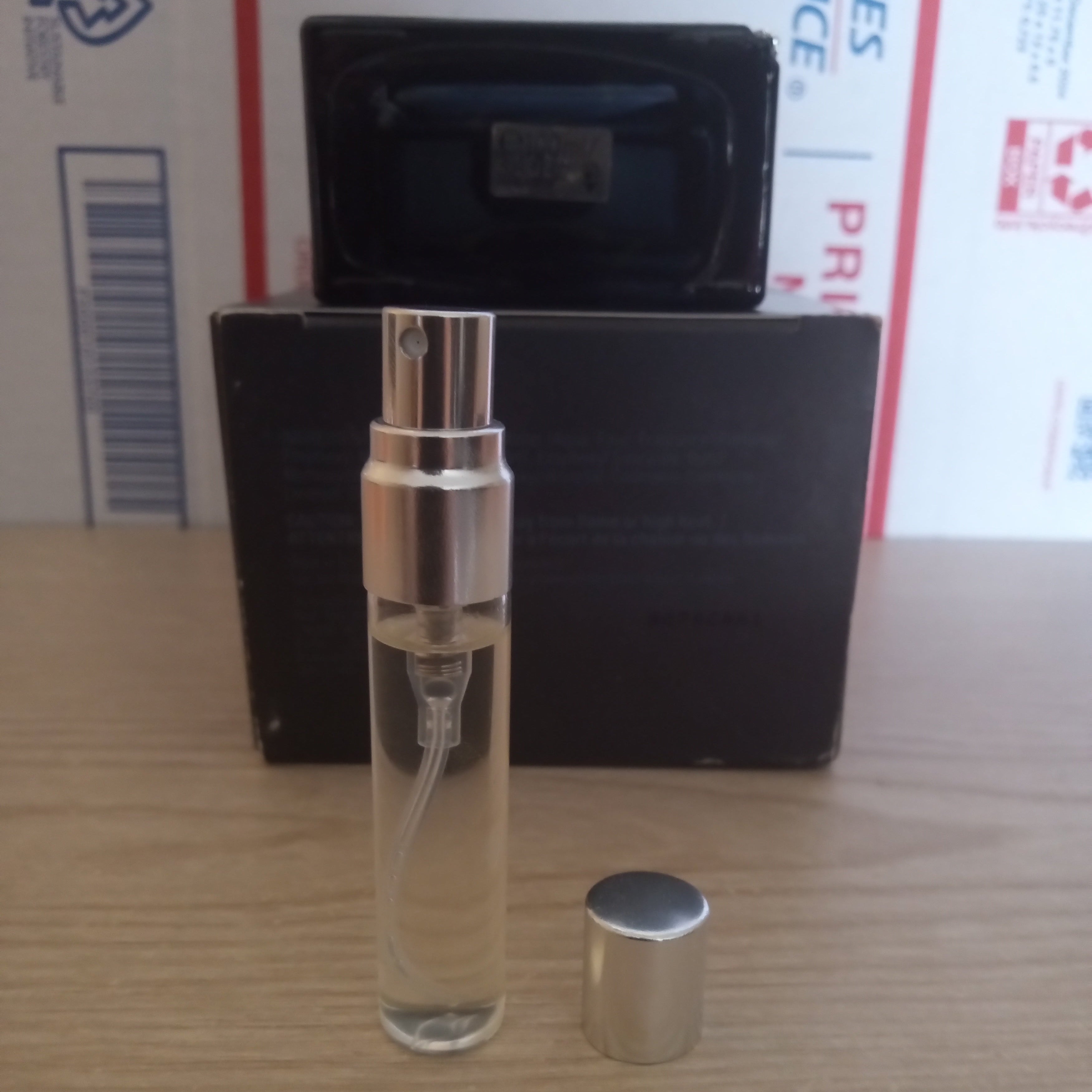 A clear glass perfume atomizer spray bottle with a silver spray nozzle, shown with its cap off next to a black rectangular box on a light wood tabletop.