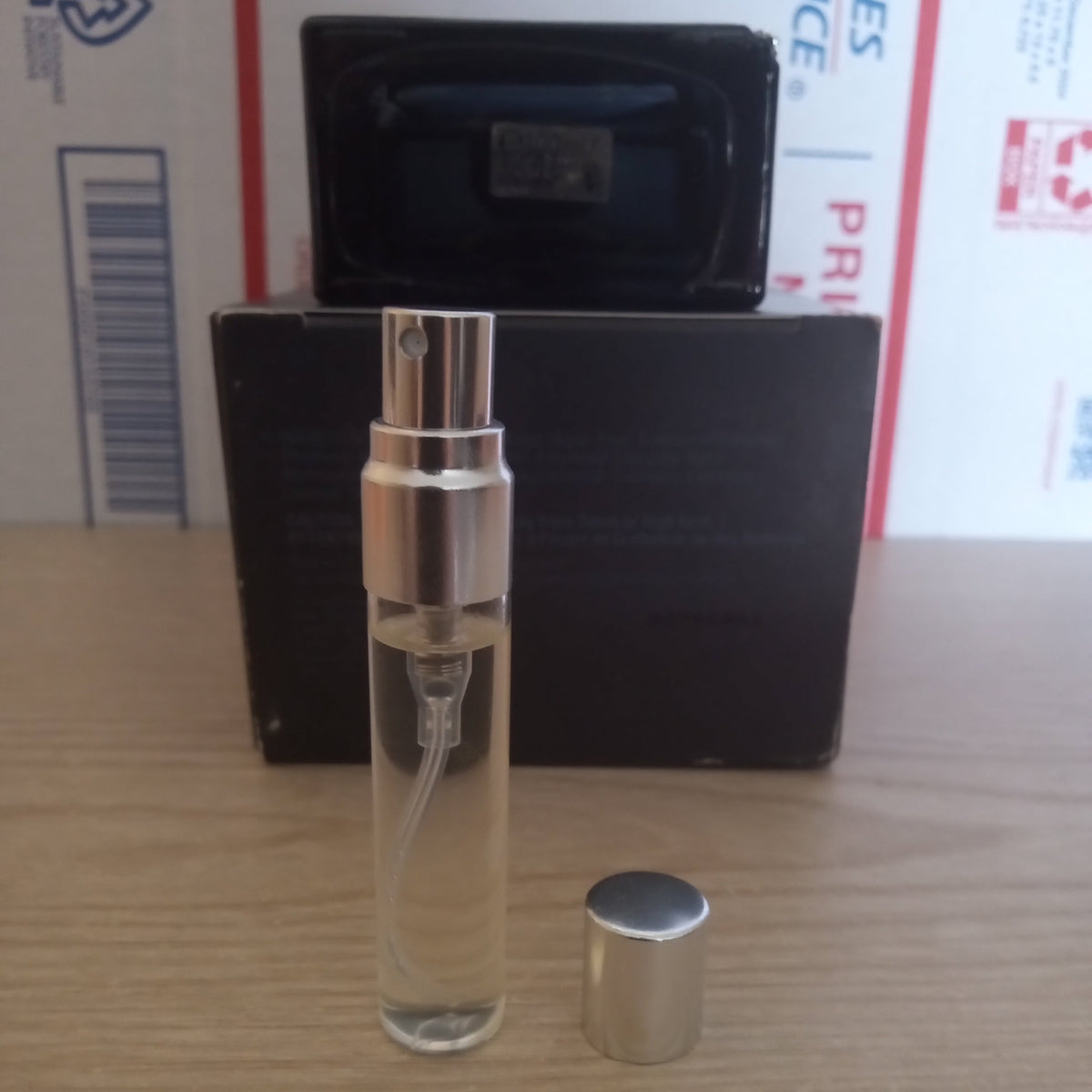 A clear glass perfume atomizer spray bottle with a silver spray nozzle, shown with its cap off next to a black rectangular box on a light wood tabletop.