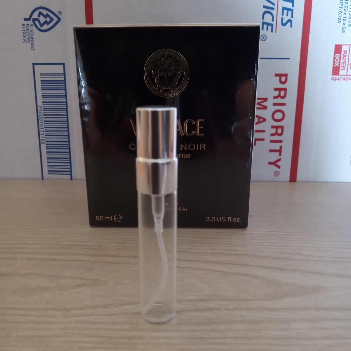 A small clear glass perfume atomizer spray vial with a silver metal collar, resting horizontally on its black cardboard packaging, which is placed on top of a USPS Priority Mail box on a light wood surface.