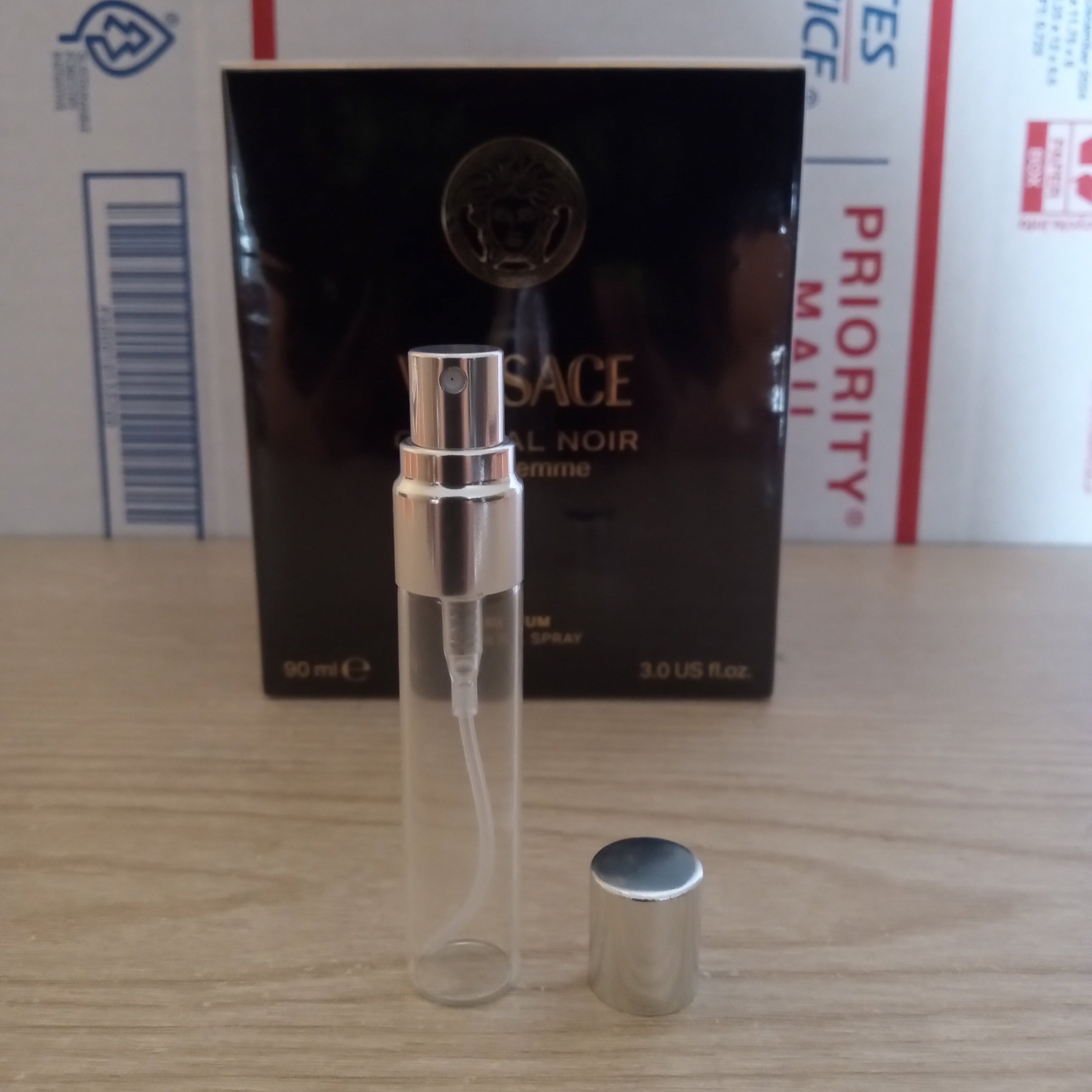 A Versace Elixir Noir 10ml clear glass perfume atomizer spray bottle with a silver cap, shown next to its black retail box on a wooden tabletop with shipping boxes in the background.